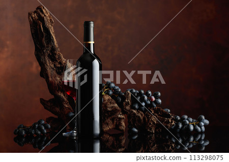Bottle and glass of red wine with an old snag and blue grapes. 113298075