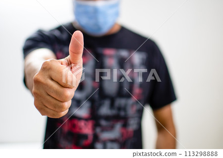 Young male with a sterile protective mask prevent coronavirus drainage or covid-19 on standing at home and showing thumbs up. Tack closeup photo and selective focus on hand. White and blur background. Young male with a sterile protective mask prevent coronavirus drainage or covid-19 on standing at home and showing thumbs up. Tack closeup photo and selective focus on hand. White and blur background. 113298888