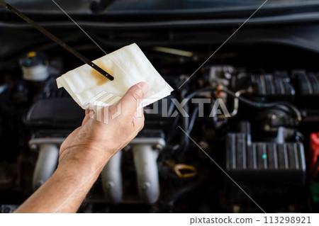 Concept of engine maintenance. A technician is checking the level of the engine oil. Man holding white tissue paper to wipe the engine oil. Close up, Blurred background Concept of engine maintenance. A technician is checking the level of the engine oil. Man holding white tissue paper to wipe the engine oil. Close up, Blurred background 113298921