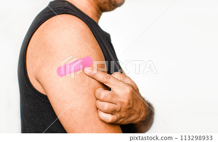 The pink plaster is attached to the man's arm. Concept for first aid after coronavirus (COVID-19) vaccination, and professional, medical, needle, blood, cancer. Closeup, white blurred background The pink plaster is attached to the man's arm. Concept for first aid after coronavirus (COVID-19) vaccination, and professional, medical, needle, blood, cancer. Closeup, white blurred background 113298933