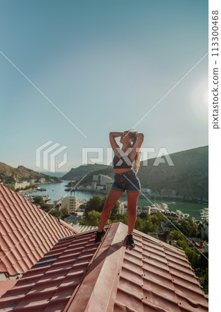 Woman standing on rooftop, enjoys town view and sea mountains. Peaceful rooftop relaxation. Below her, there is a town with several boats visible in the water. Rooftop vantage point. Woman standing on rooftop, enjoys town view and sea mountains. Peaceful rooftop relaxation. Below her, there is a town with several boats visible in the water. Rooftop vantage point. 113300468