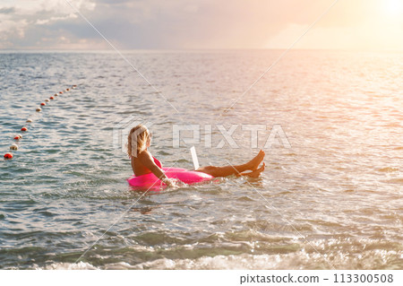 Woman works on laptop in sea. Freelancer, blond woman in sunglases floating on an inflatable big pink donut with a laptop in the sea at sunset. People summer vacation rest lifestyle concept. Woman works on laptop in sea. Freelancer, blond woman in sunglases floating on an inflatable big pink donut with a laptop in the sea at sunset. People summer vacation rest lifestyle concept. 113300508