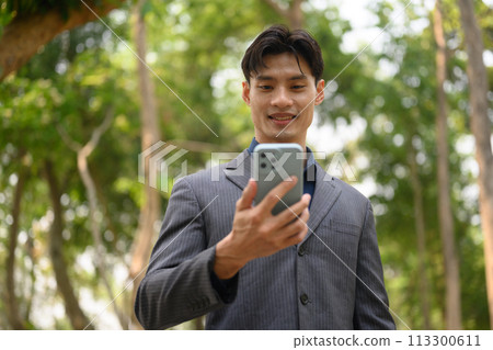 Confident young businessman text messaging on smart phone standing in city park 113300611