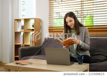 Smiling caucasian woman reading notes and working with laptop in living room 113300615