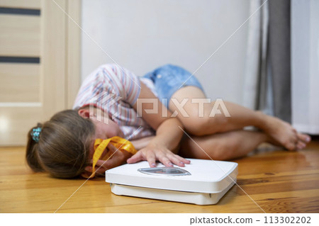 Distressed girl lying on the floor next to a weighing scale and measuring tape Distressed girl lying on the floor next to a weighing scale and measuring tape 113302202