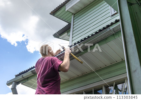 Adult man, owner of dacha with nail and an axe replacing hammer examines roof for repair. Reparation with his own hands. Rear view 113303845