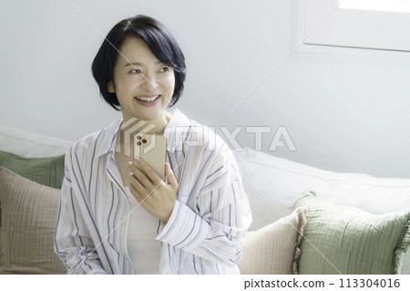 A middle-aged woman sitting on a sofa in the living room looking at her smartphone A middle-aged woman sitting on a sofa in the living room looking at her smartphone 113304016
