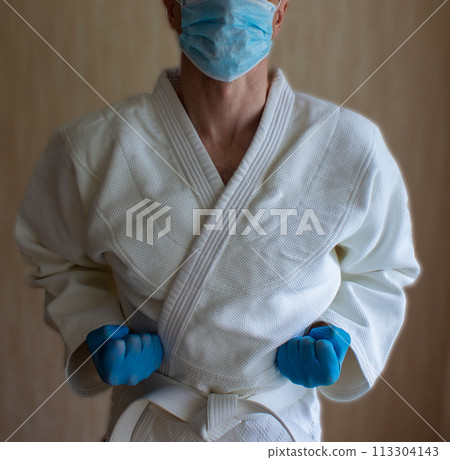 COVID-19 Pandemic Coronavirus concept. A young strong man in a white kimono for sambo, jiu jitsu and other martial arts with a blue medical gloves and medical mask. 113304143