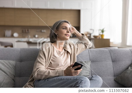 Mature woman hold cellphone sitting on couch looking away 113307082