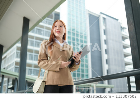 A young Asian businesswomen wearing a suit holding files standing in a big city on a busy downtown street. Young Asian businesswoman using smartphone texting to contact clients. 113307502