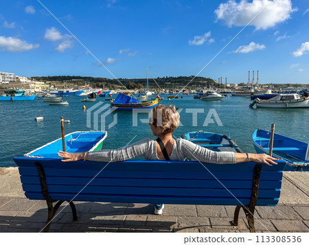 Woman sitting on bench and looking at boats in Marsaxlokk harbor, Malta 113308536