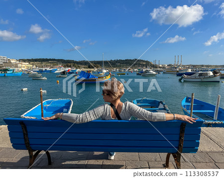 Woman sitting on bench and looking at boats in Marsaxlokk harbor, Malta 113308537