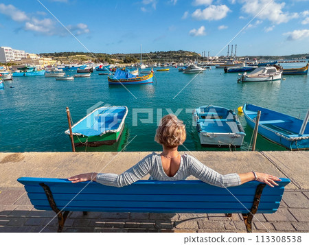 Woman sitting on bench and looking at boats in Marsaxlokk harbor, Malta Woman sitting on bench and looking at boats in Marsaxlokk harbor, Malta 113308538