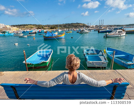 Woman sitting on bench and looking at boats in Marsaxlokk harbor, Malta Woman sitting on bench and looking at boats in Marsaxlokk harbor, Malta 113308539