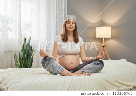 Caucasian brown haired pregnant woman meditating while sitting on bed in lotus pose practicing yoga for relaxing and calming down wearing pajama and sleeping mask Caucasian brown haired pregnant woman meditating while sitting on bed in lotus pose practicing yoga for relaxing and calming down wearing pajama and sleeping mask 113308705
