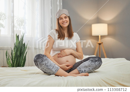 Attractive smiling cheerful Caucasian pregnant woman sitting in bed and holding her belly looking at camera with happy smile enjoying her pregnancy stroking tummy Attractive smiling cheerful Caucasian pregnant woman sitting in bed and holding her belly looking at camera with happy smile enjoying her pregnancy stroking tummy 113308712