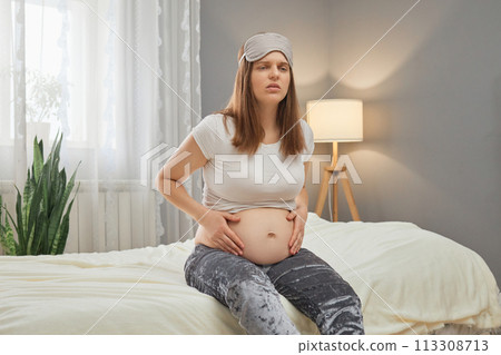 Unhealthy pregnant woman holding her belly while sitting on bed at home suffering from spasms starting contractions grimacing from pain wearing pajama and sleeping mask Unhealthy pregnant woman holding her belly while sitting on bed at home suffering from spasms starting contractions grimacing from pain wearing pajama and sleeping mask 113308713