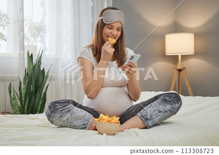 Attractive beautiful pregnant woman using mobile phone and eating potato chips while sitting on bed in her bedroom expectant mother holding phone and bowl with fast food snack Attractive beautiful pregnant woman using mobile phone and eating potato chips while sitting on bed in her bedroom expectant mother holding phone and bowl with fast food snack 113308723