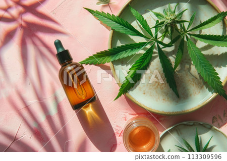Hemp cosmetic oil in dropper bottle, plate with hemp flowers and leaves on light pink pastel background. Aesthetic background with medicinal marijuana in beauty industry 113309596
