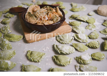 A clay plate with freshly cooked dumplings with mashed potatoes with steam, on the background of floured marble table with raw Ukrainian Varenyky. Culinary. Homemade cooking. Food background 113309632