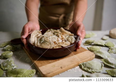 Close-up of the hands of housewife serving delicious homemade dumplings in a clay dish, standing at floured marble kitchen table with raw uncooked varennyki. Homemade food concept. Ethnic culinary Close-up of the hands of housewife serving delicious homemade dumplings in a clay dish, standing at floured marble kitchen table with raw uncooked varennyki. Homemade food concept. Ethnic culinary 113309633