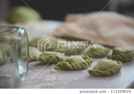 Close-up homemade dumplings of green color on a floured kitchen table and partial view of a glass bottle as rolling pinon the foreground. Traditional cuisine in Ukraine - varennyky 113309639