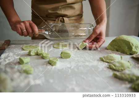Close-up hands of woman in beige apron, using a glass wine bottle for rolling out the dough with green spinach, cooking traditional Ukrainian Varennyky with mashed potatoes. Food. Traditions. Culture 113310933