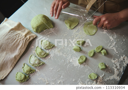 Top view woman's hands using a glass wine bottle for rolling out the dough with green spinach, cooking traditional Ukrainian Varennyky with mashed potatoes. Food. Traditions. Culture 113310934