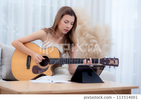 Young asian woman sitting on sofa looking tablet for practice music with guitar in living room. Young asian woman sitting on sofa looking tablet for practice music with guitar in living room. 113314001