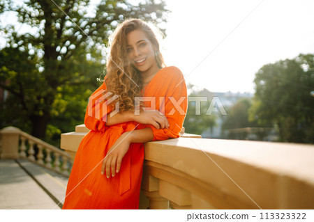 Young woman posing against backdrop of urban landscape in bright orange dress. Concept of lifestyle. Young woman posing against backdrop of urban landscape in bright orange dress. Concept of lifestyle. 113323322