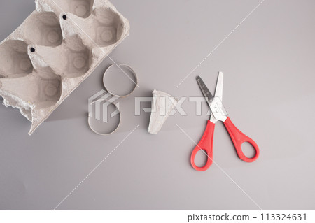Minimalistic DIY project scene: Empty egg carton placed on a grey table. Disassembled paper hoop, part of the cardboard-based tutorial for crafting  113324631