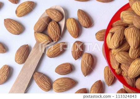 Close-up shot of almonds in a red bowl and wooden spoon isolated on white background. Close-up shot of almonds in a red bowl and wooden spoon isolated on white background. 113324843