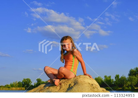 Adorable little girl sitting on a big stone in the lake on a hot summer day. Child dressed in swimsuit. Summer and happy childhood concept. Copy space in bright blue sky Adorable little girl sitting on a big stone in the lake on a hot summer day. Child dressed in swimsuit. Summer and happy childhood concept. Copy space in bright blue sky 113324953