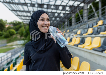 A Muslim woman in hijab holding a water bottle at a sports stadium, embodying health and fitness. A Muslim woman in hijab holding a water bottle at a sports stadium, embodying health and fitness. 113326571