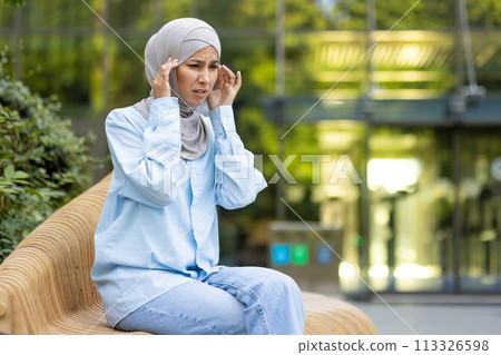 A woman wearing a hijab outdoors appears stressed and holds her head, showing signs of a headache or anxiety. 113326598
