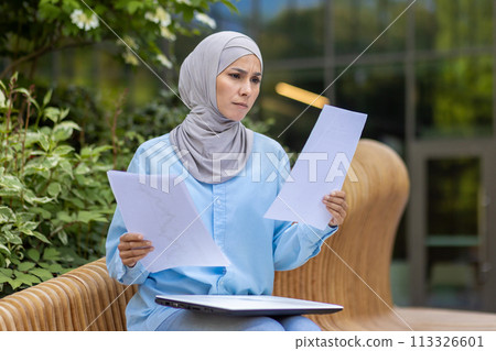 A focused professional woman in a hijab examines papers, sitting on an outdoor bench in a tranquil setting. 113326601