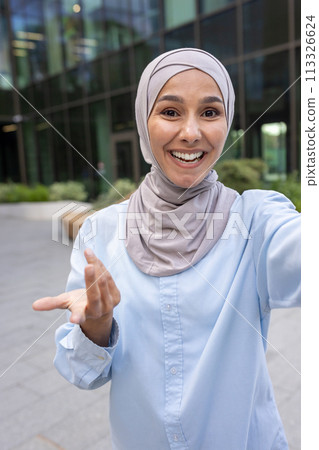 Cheerful Muslim woman with hijab captures a selfie with a smartphone, smiling joyfully in a modern city environment. Cheerful Muslim woman with hijab captures a selfie with a smartphone, smiling joyfully in a modern city environment. 113326624