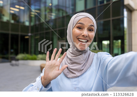 Cheerful woman in a hijab capturing a selfie moment outdoors with a high five gesture and joyful expression. 113326628