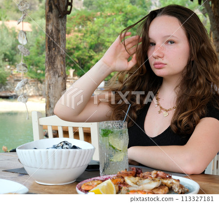 Young woman in a restaurant mojito and seafood on a plate against the backdrop of green river in Albania delicious food cooking relax straightening her hair black dress loose brown hair. High quality Young woman in a restaurant mojito and seafood on a plate against the backdrop of green river in Albania delicious food cooking relax straightening her hair black dress loose brown hair. High quality 113327181