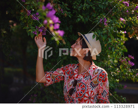young asian woman stands in sunny green park, admiring blooming bougainvillea bush. The flower's bright colors are beautiful contrast to park's lush greenery. young asian woman stands in sunny green park, admiring blooming bougainvillea bush. The flower's bright colors are beautiful contrast to park's lush greenery. 113328971