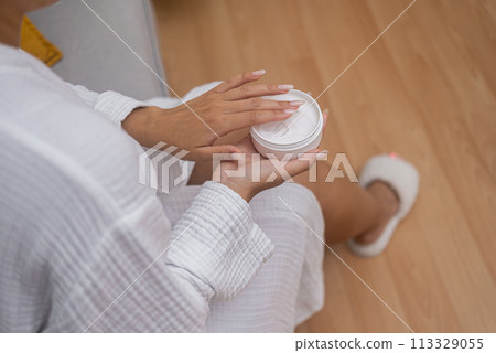 Home body care: A woman in a bathrobe hydrates her skin using a moisturizing lotion from a jar, perfecting her skincare regimen.  113329055