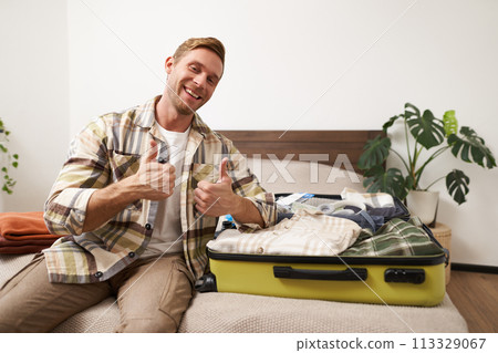 Portrait of young handsome man, tourist going on vacation, packing luggage for holidays, putting clothes in suitcase, sitting on bed 113329067