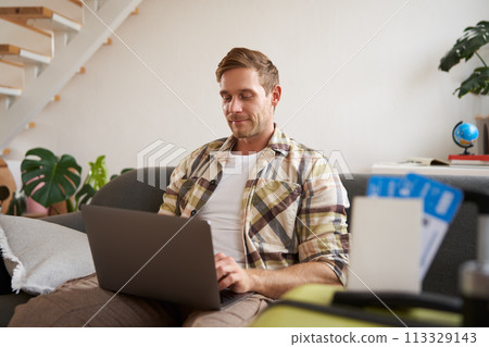 Portrait of handsome young man, has two tickets and passport, goes online on laptop, booking hotel and planning his holiday, sitting in living room Portrait of handsome young man, has two tickets and passport, goes online on laptop, booking hotel and planning his holiday, sitting in living room 113329143