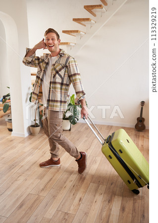 Full length photo of happy guy with suitcase, walking in apartment, shows phone call gesture Full length photo of happy guy with suitcase, walking in apartment, shows phone call gesture 113329219