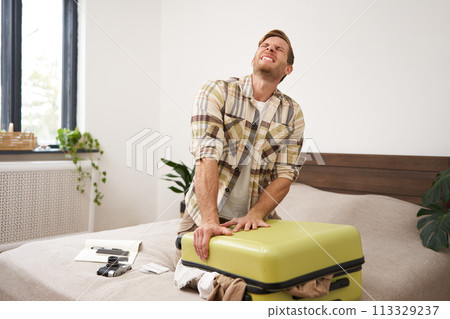 Portrait of young man sweating, shutting his suitcase, trying to close the luggage with lots of clothes, sitting on bed, packing things for summer vacation, going on holiday or business trip Portrait of young man sweating, shutting his suitcase, trying to close the luggage with lots of clothes, sitting on bed, packing things for summer vacation, going on holiday or business trip 113329237