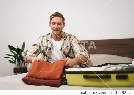 Portrait of handsome young man, tourist unpacking clothes in hotel room, staying in hostel on vacation, travelling, sitting with luggage on bed 113329262