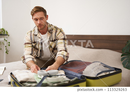 Portrait of young handsome bearded guy, tourist going on holiday, fastening his clothes in suitcase, packing for vacation, going on business trip, sitting in bedroom 113329263