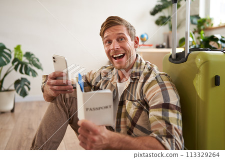 Portrait of handsome smiling man with suitcase, looks at smartphone, holds travel tickets and passport 113329264
