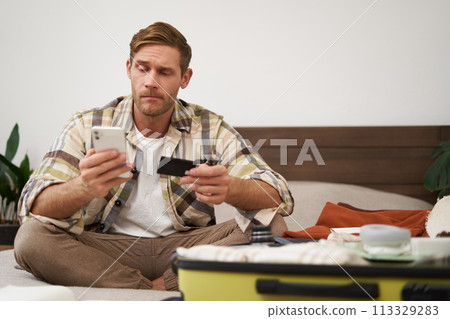 Angry man holds credit card and mobile phone, sits next to clothes in suitcase, has problems with paying online Angry man holds credit card and mobile phone, sits next to clothes in suitcase, has problems with paying online 113329283