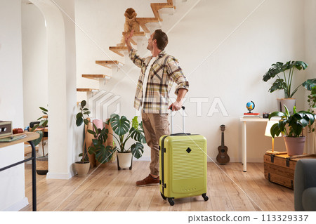 Portrait of happy traveller, young man going on vacation, holding his suitcase, petting a cat on stairs 113329337